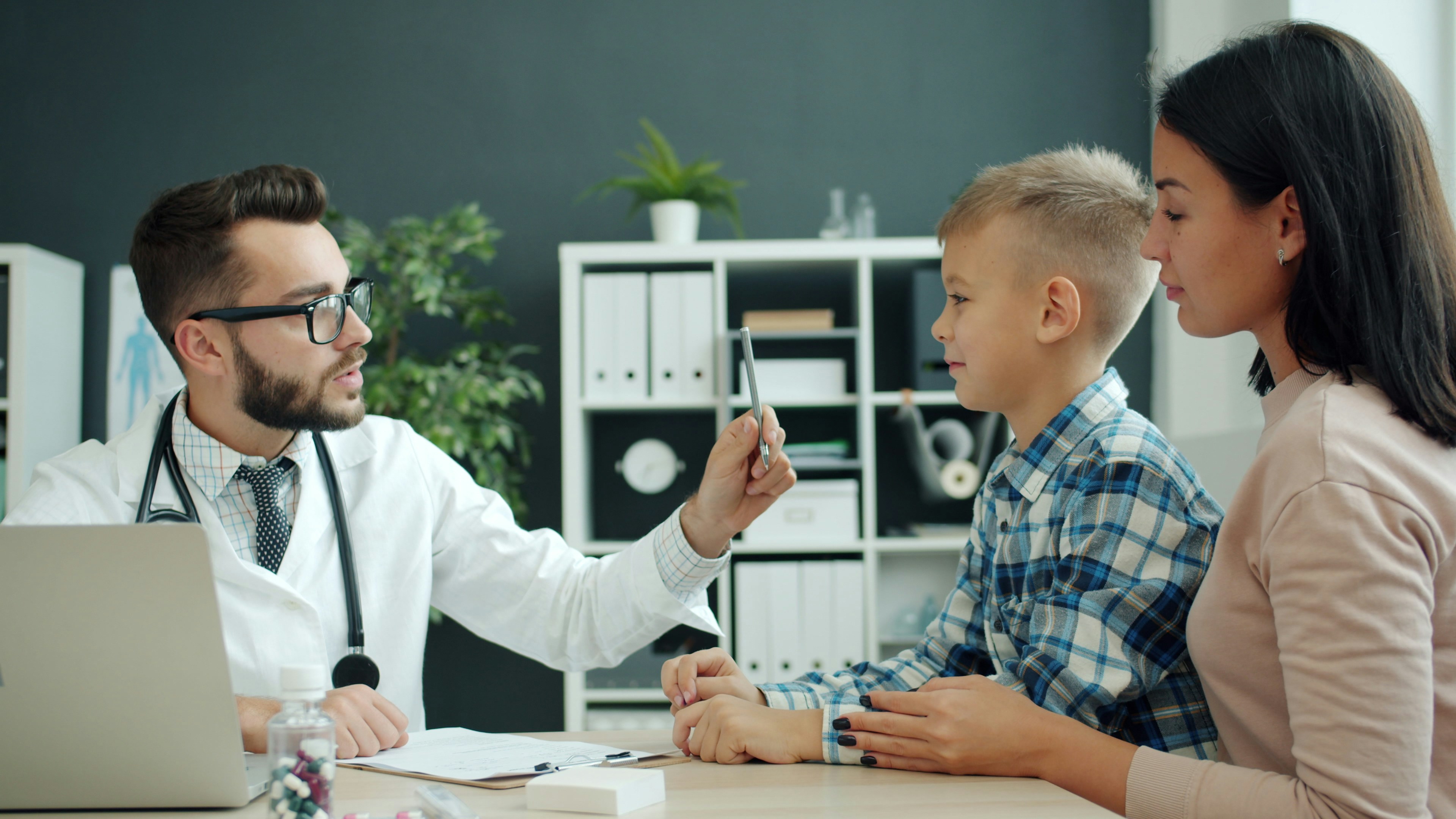 🎬 FULL 4K VIDEO for this still is in my bio link! ⬇️ Male pediatrician in white gown is talking to family young mother and son examining child writing in chart during medical appointment in hospital.